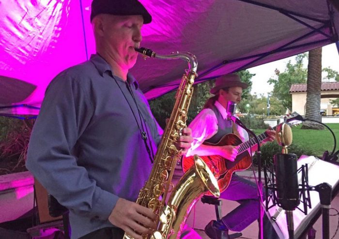 man plays saxaphone and another man behind him plays guitar at an outdoor concert. A purple light shines on them.